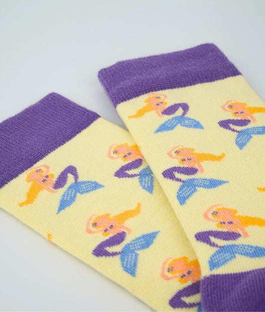 Chaussettes fantaisie jaunes et violettes en coton biologique à motif de sirène. Fabriquées en Europe, elles offrent un confort optimal grâce à leurs zones renforcées et leurs finitions plates sans couture.
