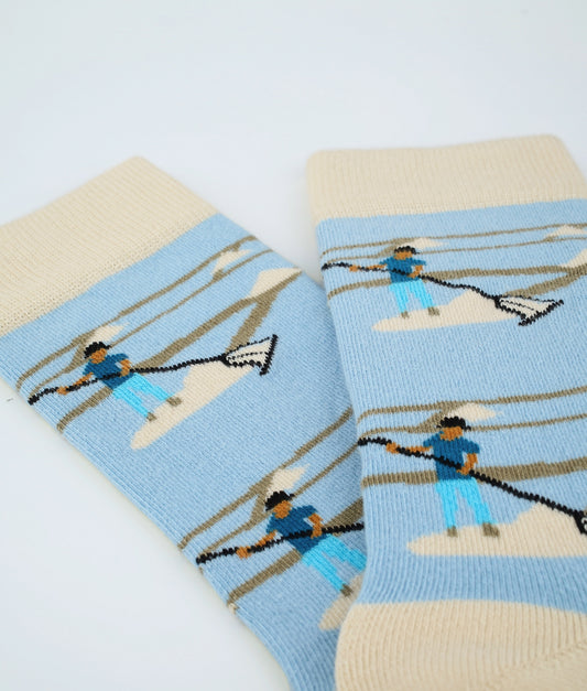 Chaussettes fantaisie bleu clair et beige à motif de sauniers. Fabriquées en Europe en coton biologique, elles offrent confort, douceur et un clin d’œil au savoir-faire local.
